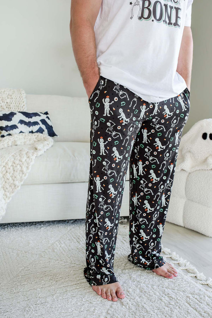 Adult wearing Caden Lane black Halloween pajama pants with white skeleton, mummy, and candy corn print, paired with a white 'Bag of Bones' graphic tee — part of Caden Lane's matching family Halloween pajama collection