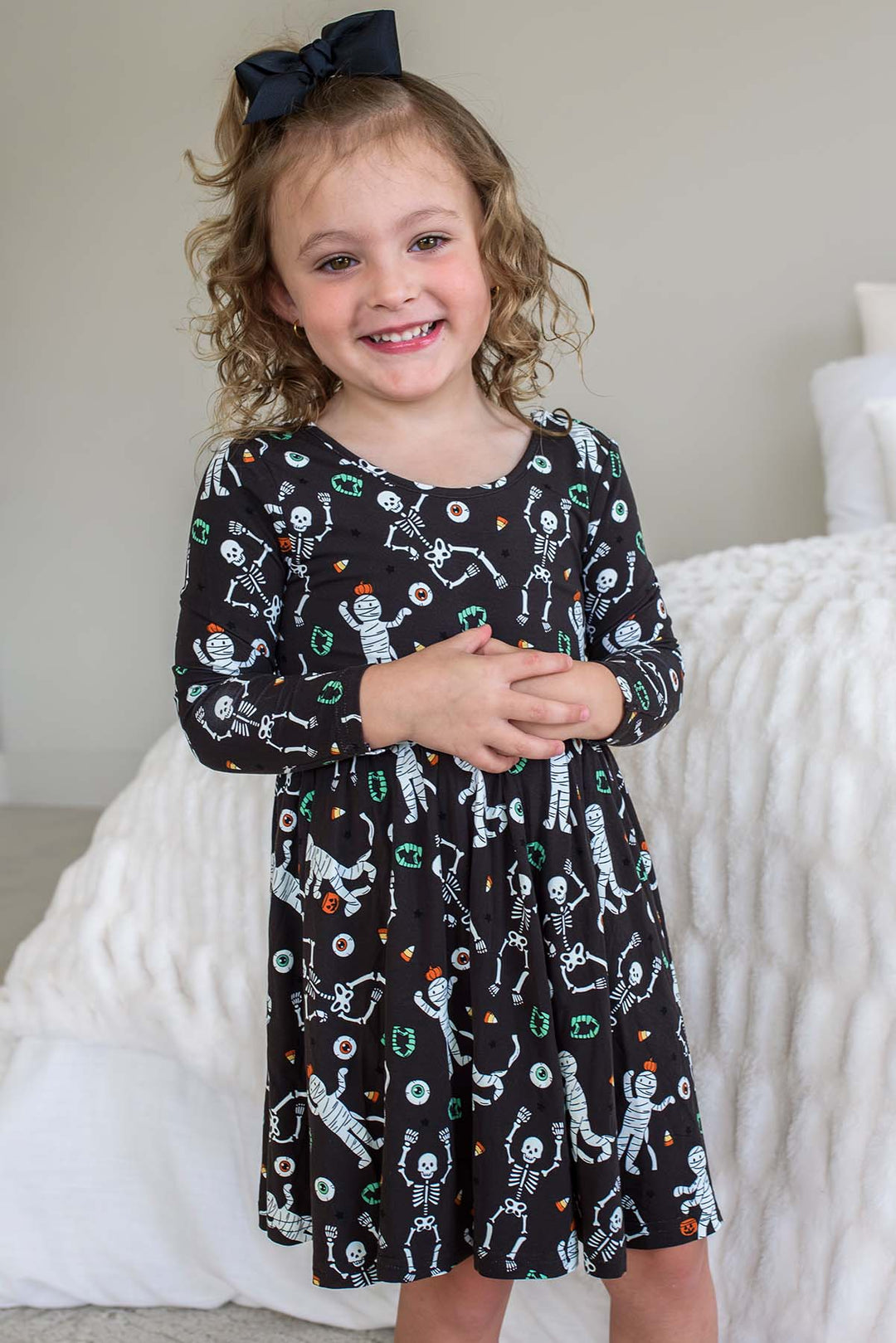 Young girl wearing a Caden Lane black long-sleeve Halloween dress with a twirl skirt, featuring a white skeleton, mummy, and candy corn print, styled with a black hair bow