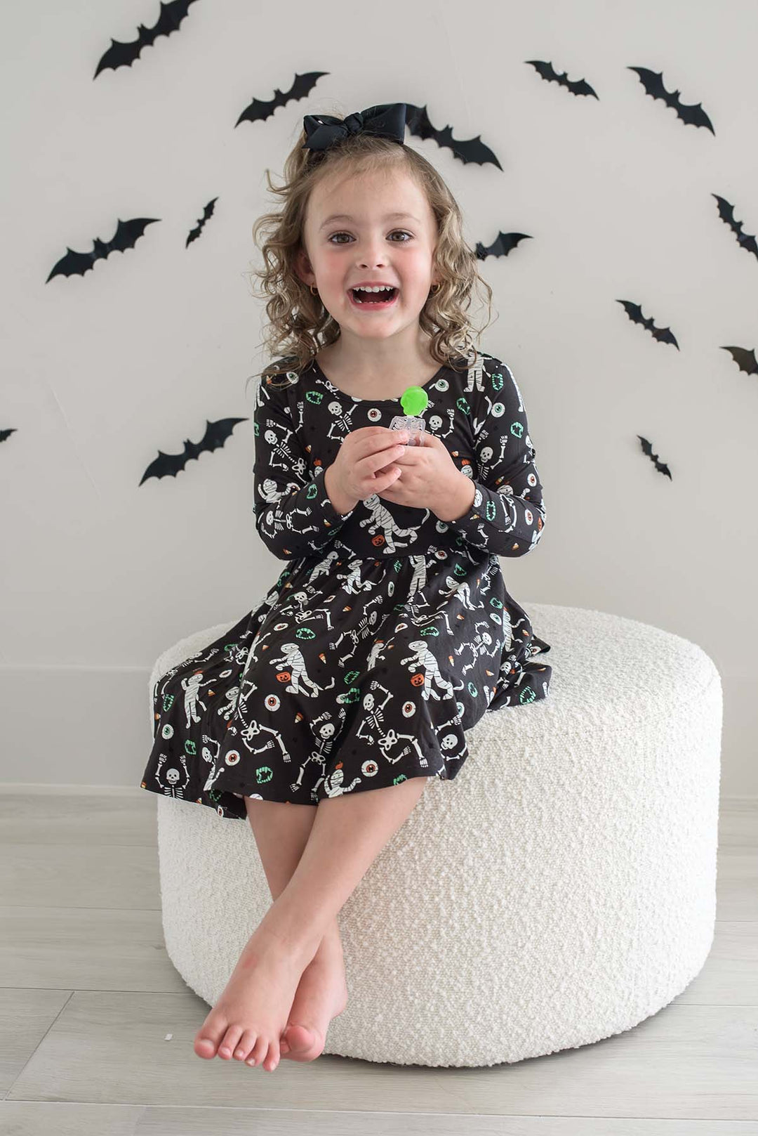 Young girl wearing a Caden Lane black long-sleeve Halloween dress with a twirl skirt, featuring a white skeleton, mummy, and candy corn print, styled with a black hair bow. With bat cutouts on white wall behind her.