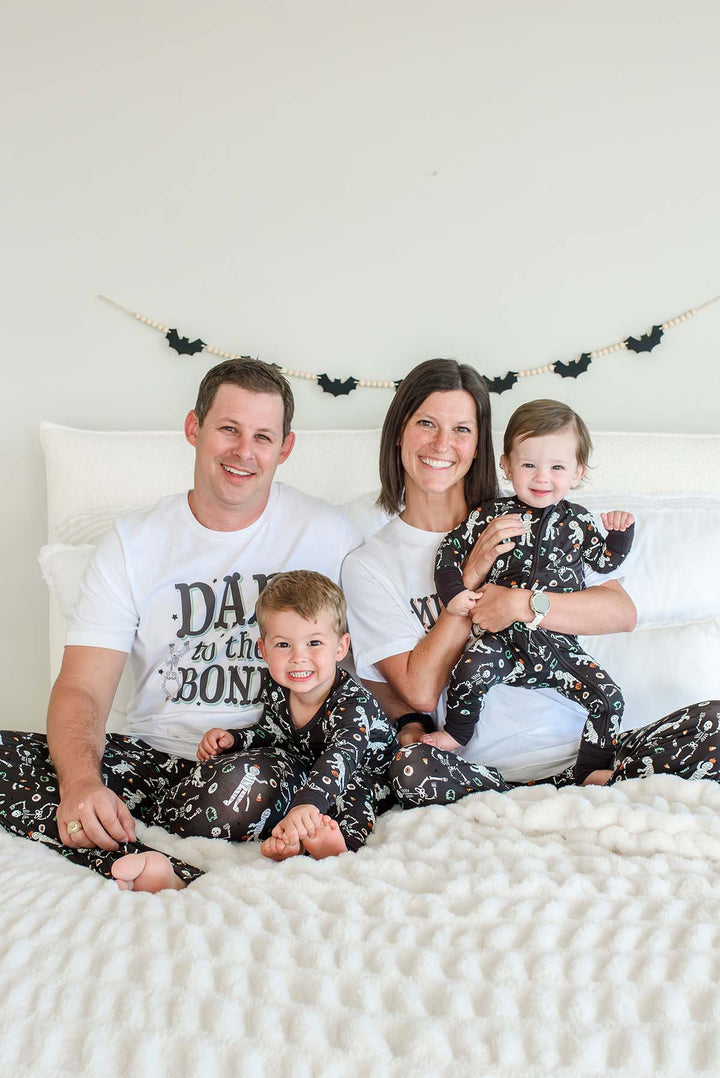 A family of four wearing matching black and white skeleton-print pajamas, sitting together on a bed with white bedding and a white headboard. The father and mother wear white graphic T-shirts, and the two children wear matching black pajamas with white skeleton prints.