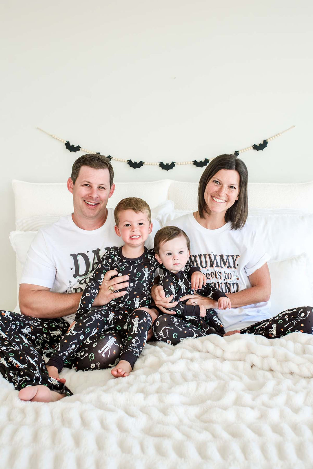 A family of four and their dog wearing matching black and white skeleton-print pajamas, posed together on a bed with white bedding and a white wall behind them