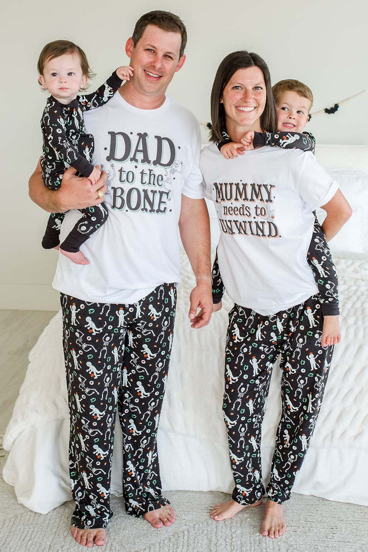 A family of four wearing matching white graphic pajama shirts and black patterned pajama pants standing in a bedroom. The dad holds a toddler on his left, and the mom holds a toddler on her right.
