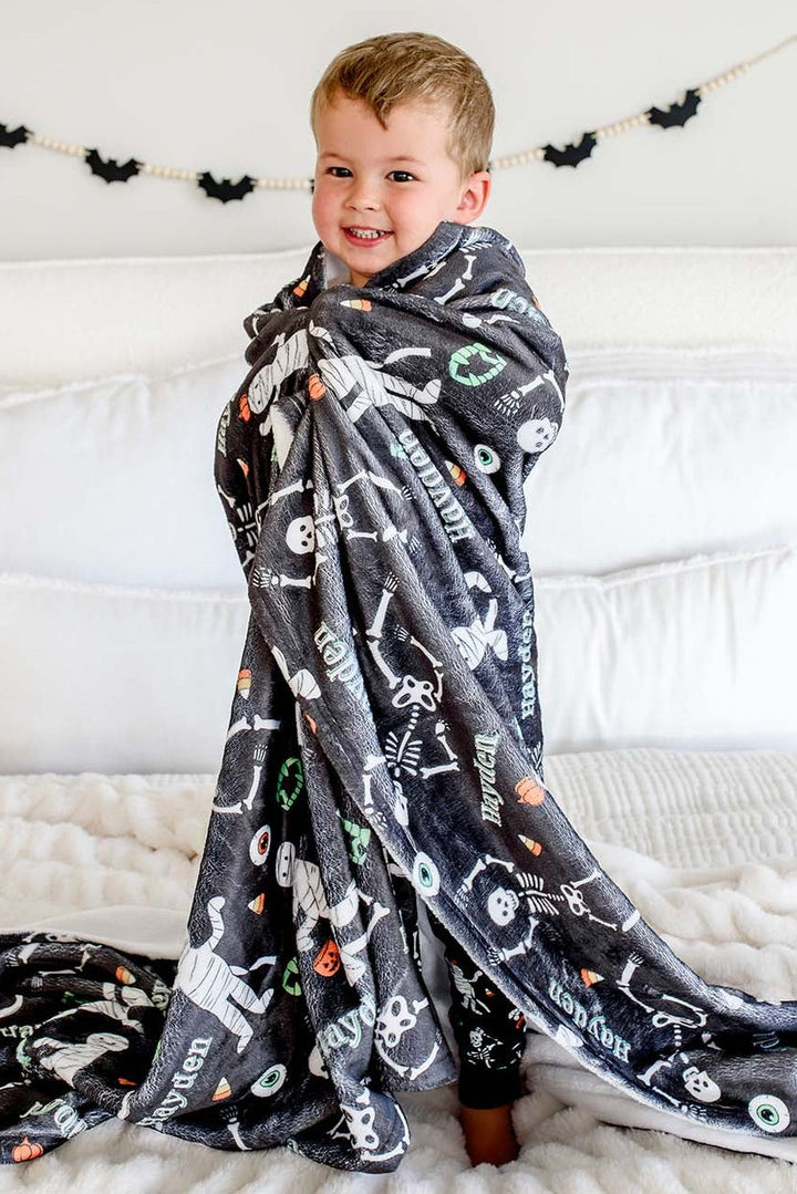 A young boy smiling while wrapped in a dark gray plush blanket with a white skeleton print, sitting on a white bed with pillows and a black bat banner hanging on the wall behind him.
