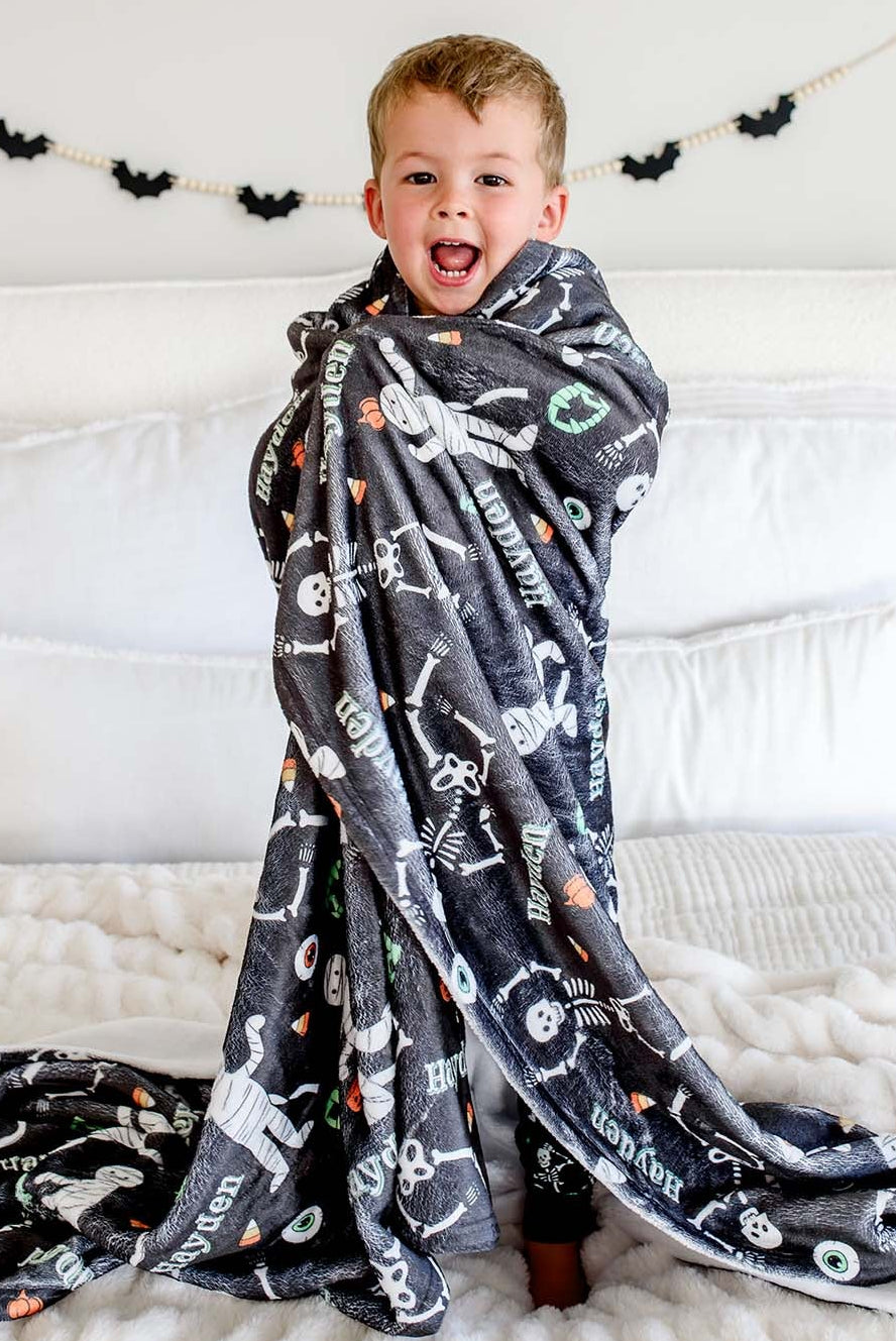 A young boy standing on a bed wrapped in a dark gray plush blanket personalized with a name, printed with white skeleton and candy corn graphics.