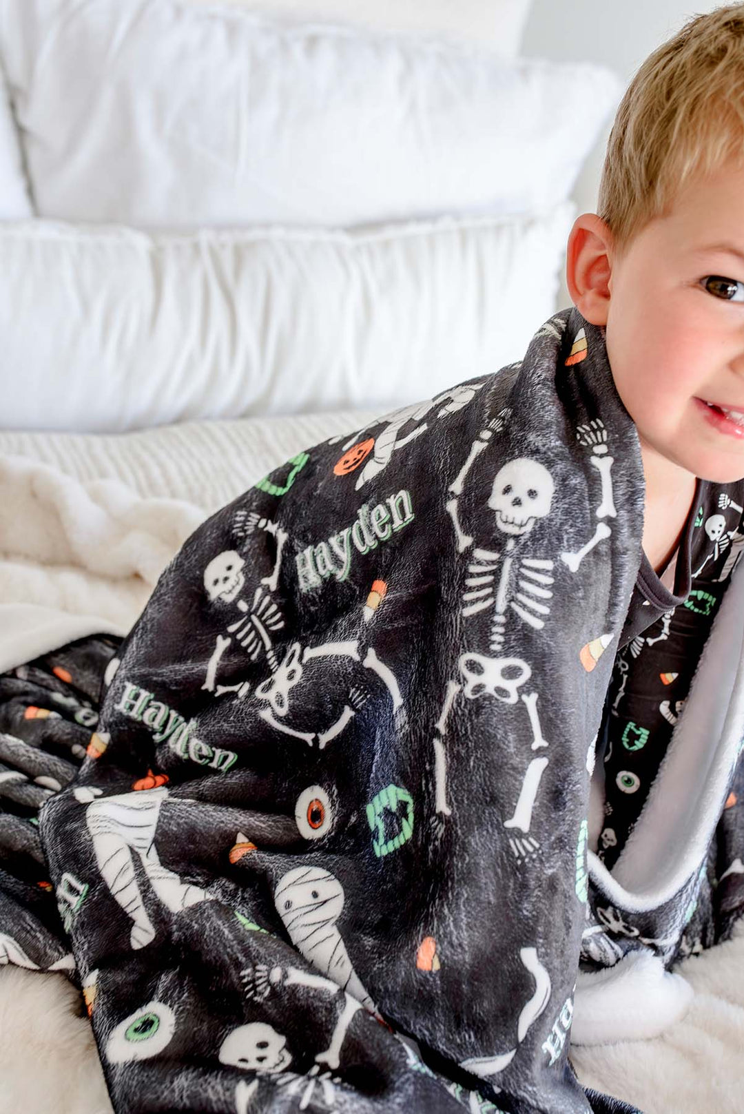 A child wearing a black plush pajama set with a white skeleton print and colorful Halloween-themed accents, wrapped in a matching black blanket with white skeletons and the name 'Hayden' printed on it, sitting on a white bed.