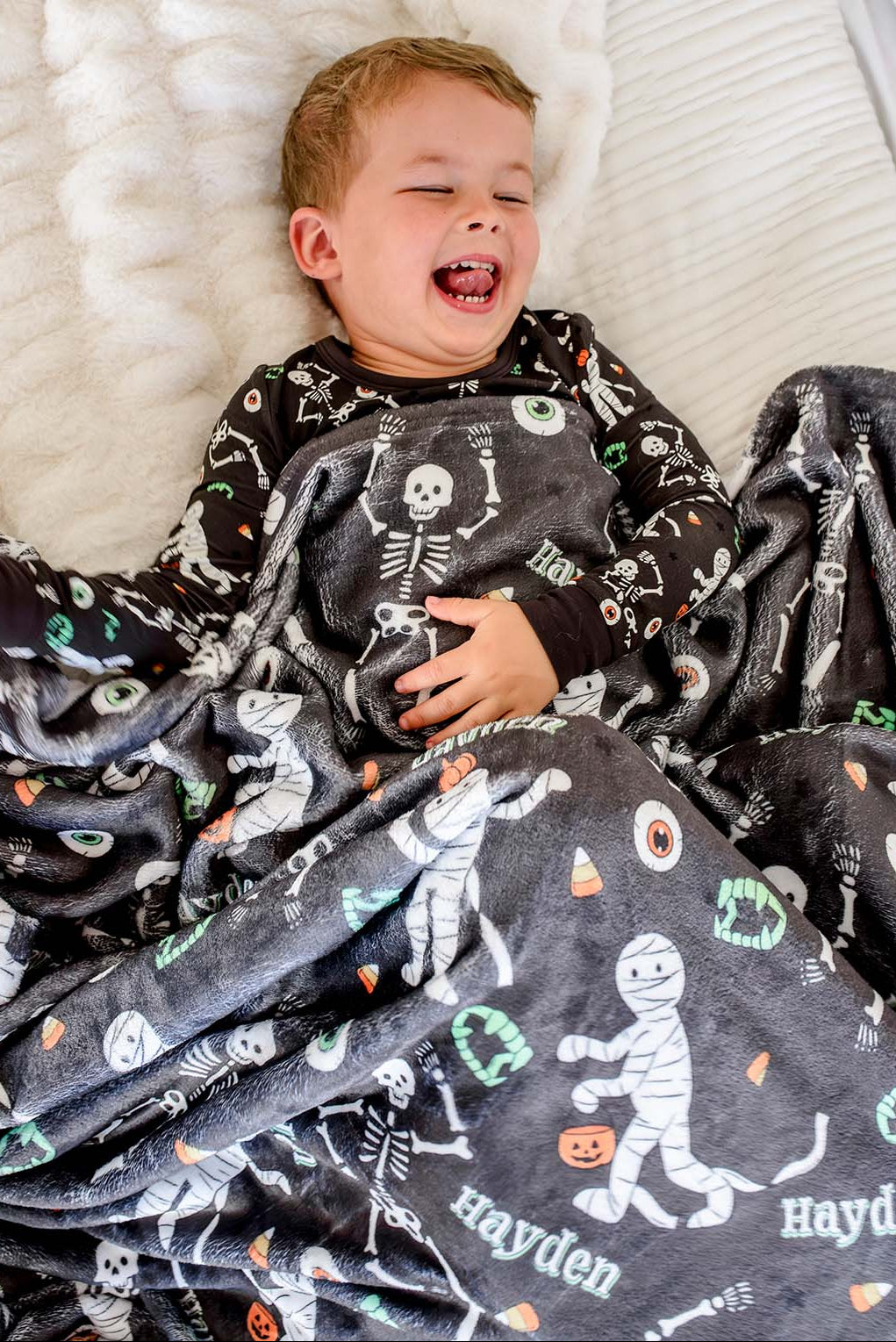 A smiling young boy lying on a white textured bed, covered with a dark gray personalized blanket featuring a white skeleton and Halloween-themed illustrations, including the name 'Hayden'.