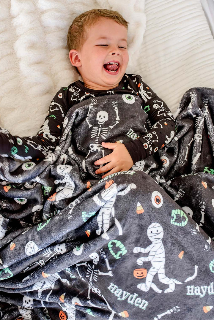 A smiling young boy lying on a white textured bed, covered with a dark gray personalized blanket featuring a white skeleton and Halloween-themed illustrations, including the name 'Hayden'.