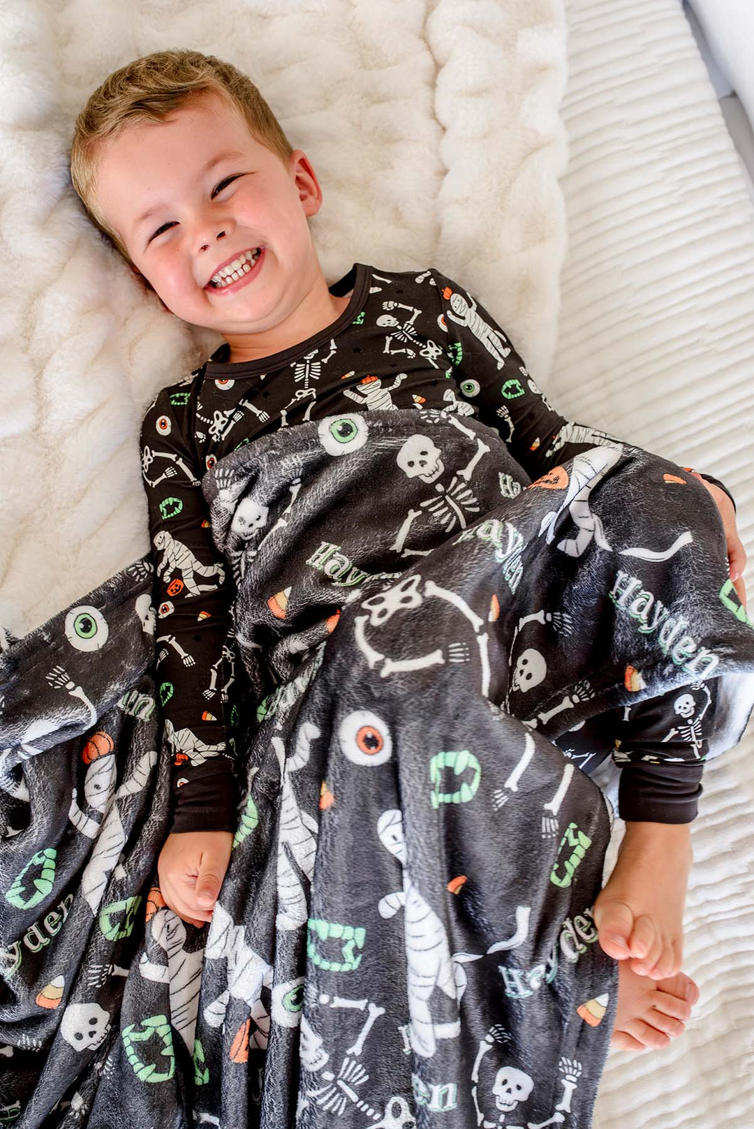 A smiling young boy lies on a white textured blanket, wearing a black long-sleeve pajama top and wrapped in a dark gray Halloween-themed blanket printed with white skeletons, mummies, eyeballs, and orange candy corn.