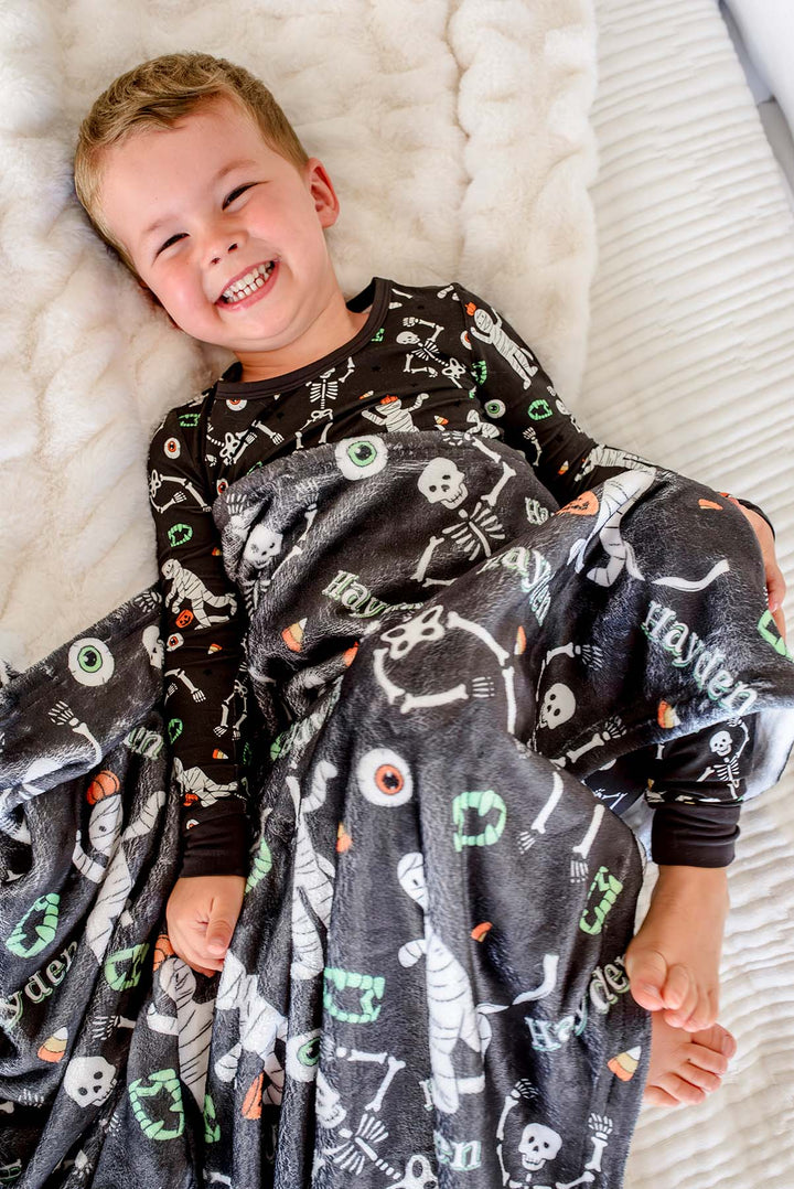 A smiling young boy lies on a white textured blanket, wearing a black long-sleeve pajama top and wrapped in a dark gray Halloween-themed blanket printed with white skeletons, mummies, eyeballs, and orange candy corn.