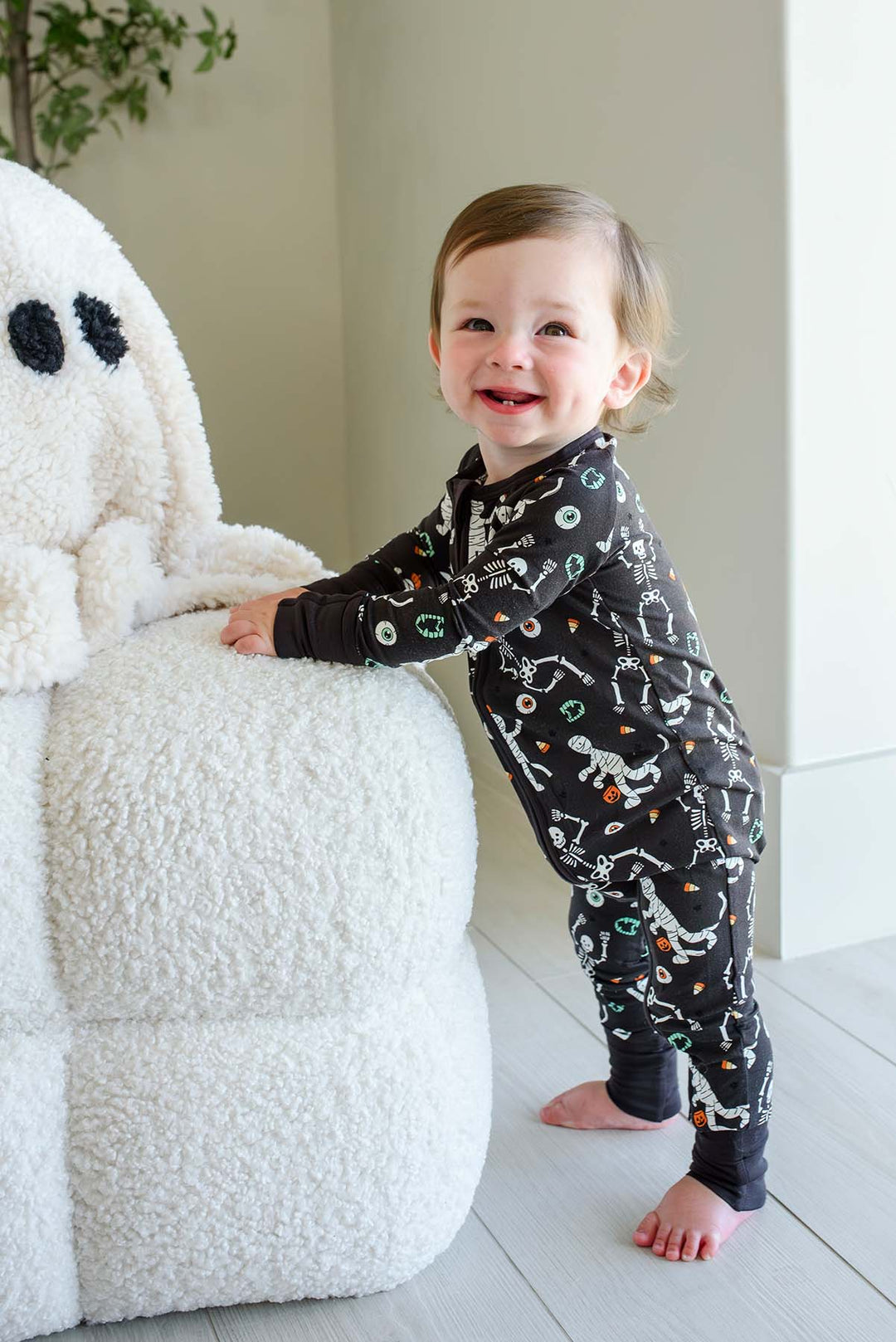 Smiling toddler standing beside a plush white ghost and holding a mummy toy, wearing a Caden Lane black zip-up skeleton and mummy print romper.