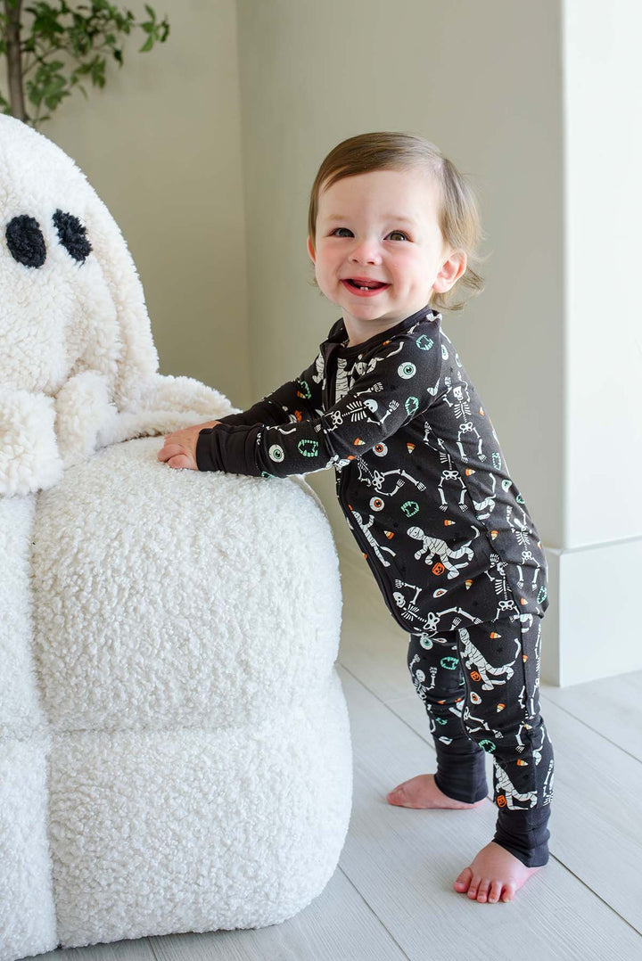 Smiling toddler standing beside a plush white ghost and holding a mummy toy, wearing a Caden Lane black zip-up skeleton and mummy print romper.