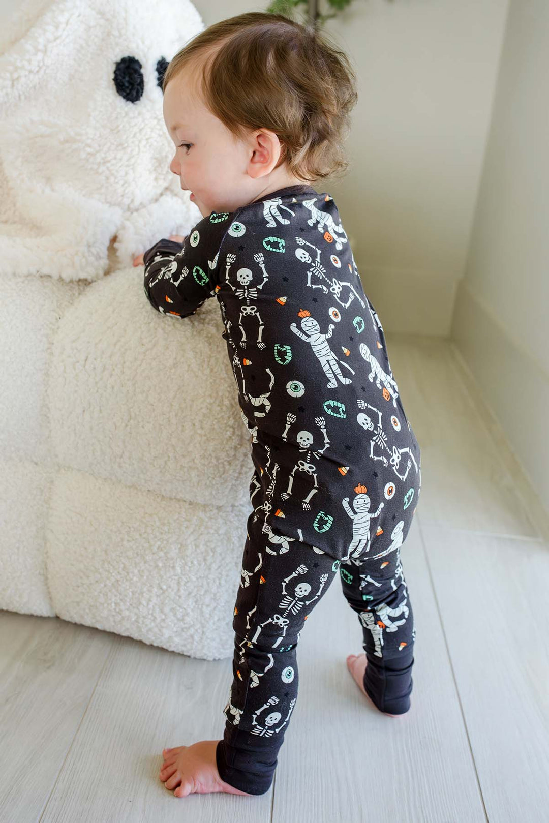 Toddler standing beside a plush white ghost and holding a mummy toy, wearing a Caden Lane black zip-up skeleton and mummy print romper.