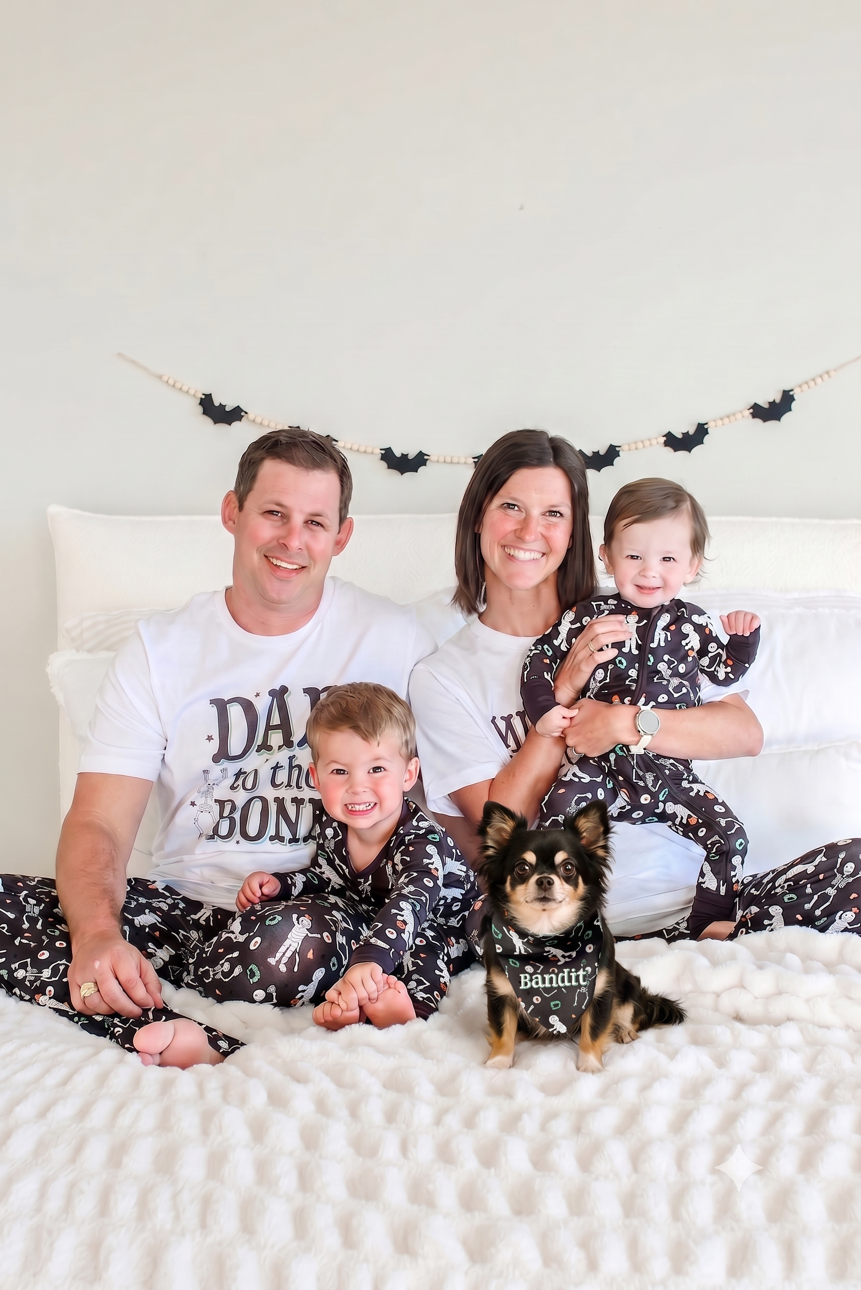 A family of four and their dog wearing matching black and white skeleton-print pajamas, posed together on a bed with white bedding and a white wall behind them.