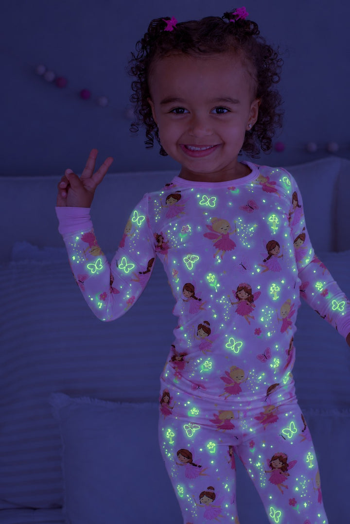 Smiling girl flashing a peace sign, wearing a glowing Caden Lane pink bamboo two-piece pajama set in a Fairy Wishes print featuring fairies, butterflies, and mushrooms.