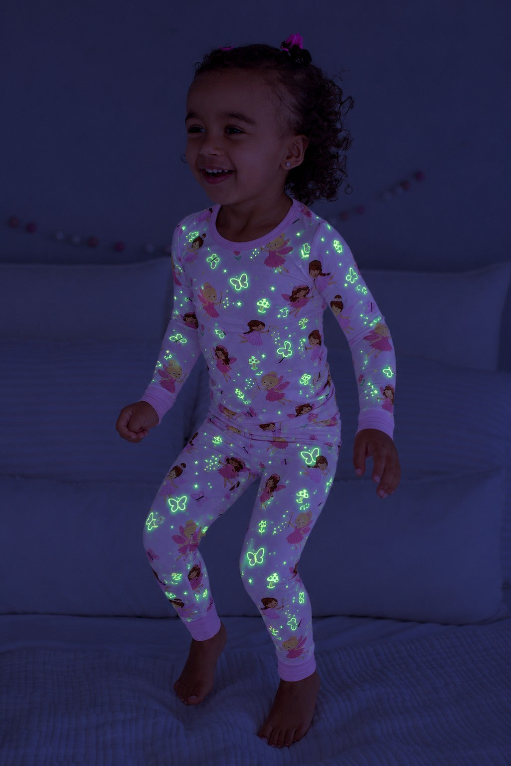 Full-length shot of a girl jumping on a bed, wearing a glowing Caden Lane pink bamboo two-piece pajama set in a Fairy Wishes print featuring fairies and butterflies.
