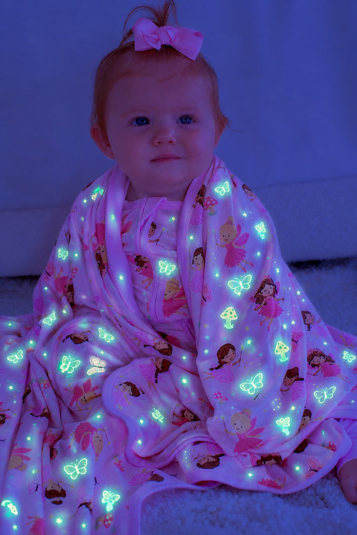 Baby with a pink hair bow sitting wrapped in a glowing Caden Lane pink bamboo blanket in a Fairy Wishes print featuring fairies, butterflies, and mushrooms.