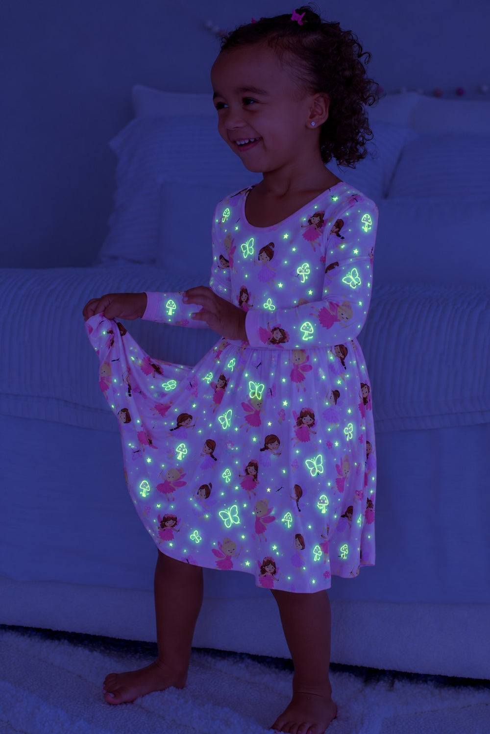 Smiling girl holding out her skirt, wearing a glowing Caden Lane pink bamboo long sleeve twirl dress in a Fairy Wishes print featuring fairies and butterflies.