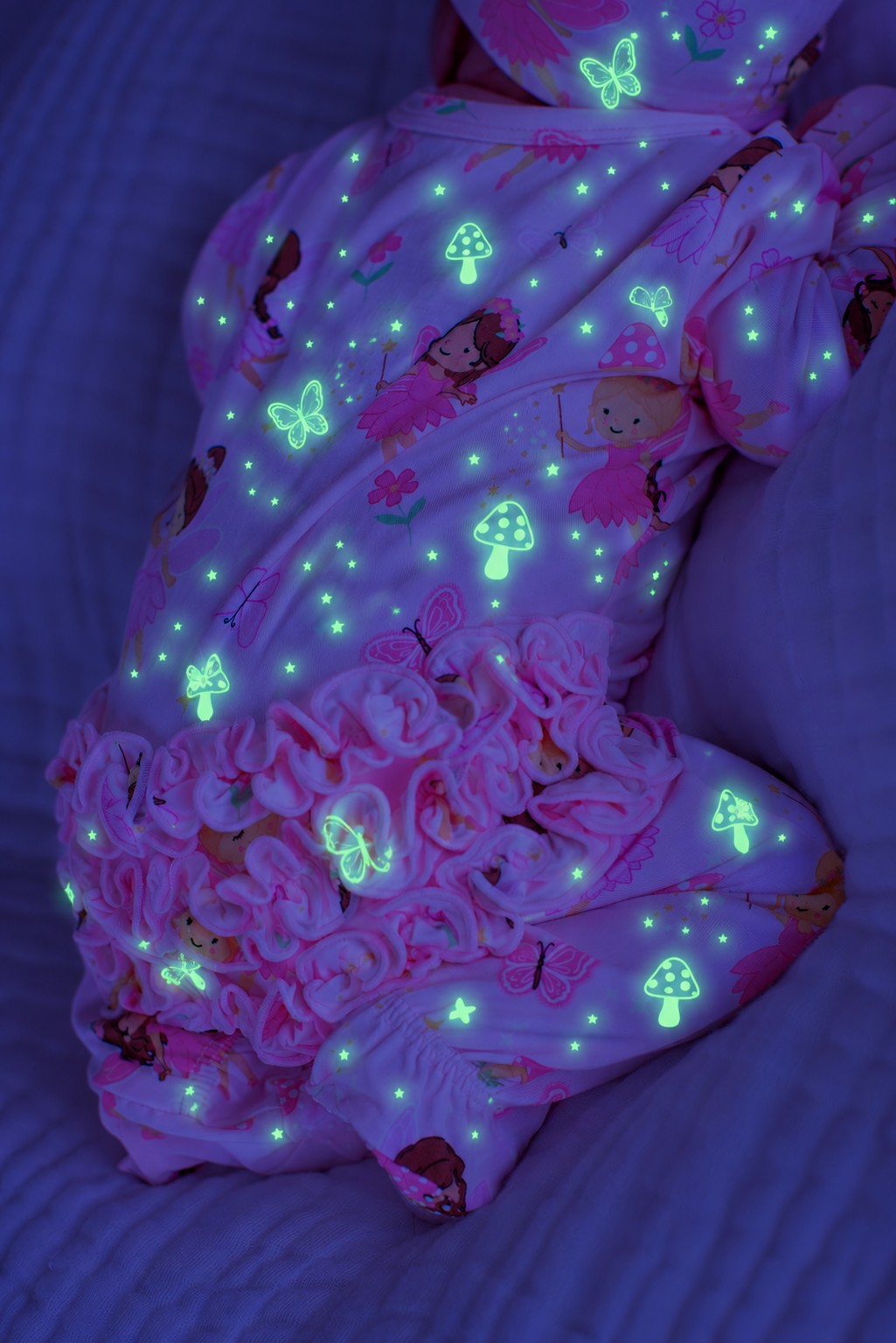 Close-up of a baby lying on a bed, wearing a glowing Caden Lane pink bamboo ruffle zipper footie in a Fairy Wishes print featuring fairies, butterflies, and mushrooms.