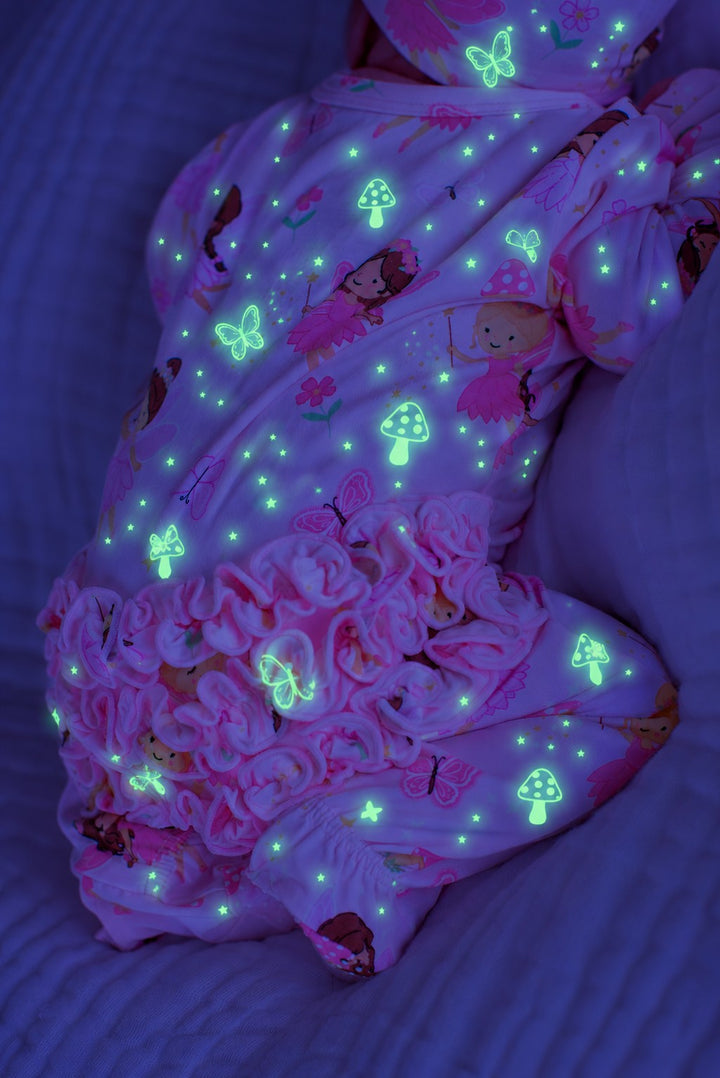 Close-up of a baby lying on a bed, wearing a glowing Caden Lane pink bamboo ruffle zipper footie in a Fairy Wishes print featuring fairies, butterflies, and mushrooms.