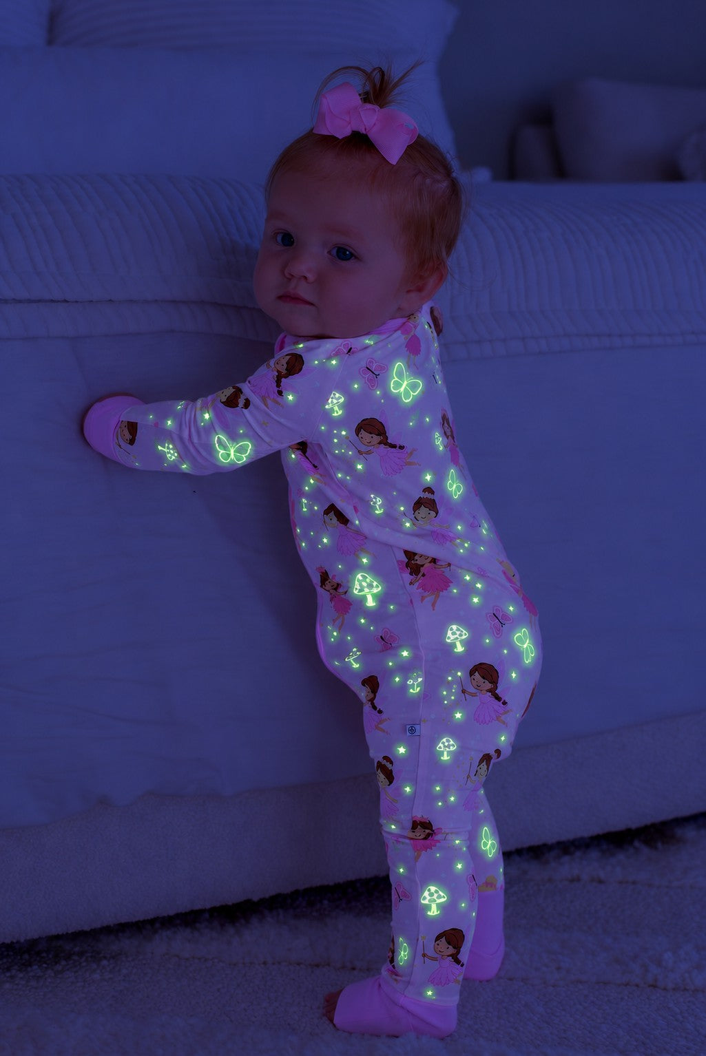 Baby with a pink hair bow standing against a couch, wearing a glowing Caden Lane pink bamboo convertible zip romper in a Fairy Wishes print featuring fairies and mushrooms.