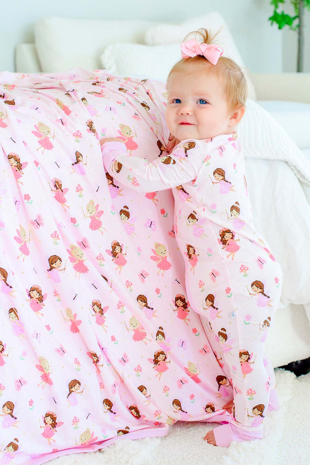 Smiling baby standing at the bed beside the Fairy Wishes glow-in-the-dark double-sided bamboo blanket, wearing matching fairy print