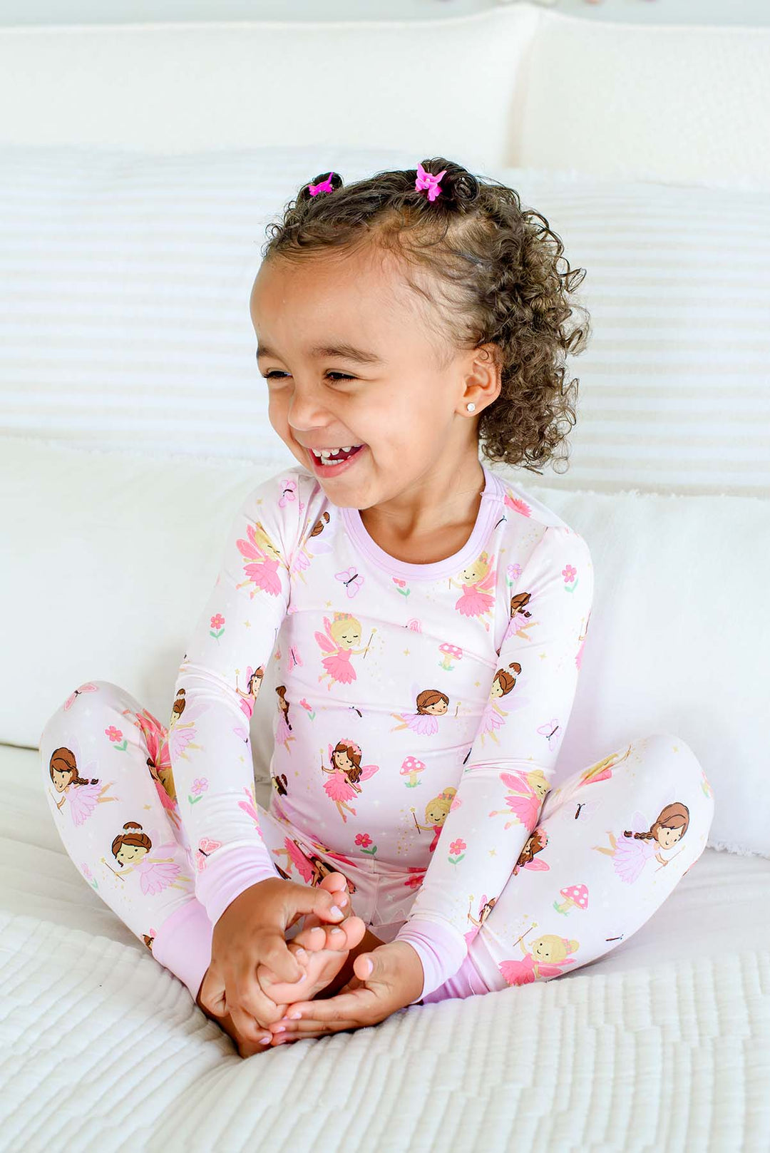 Laughing toddler girl sitting cross-legged on a bed in the Fairy Wishes glow-in-the-dark two-piece bamboo pajama set