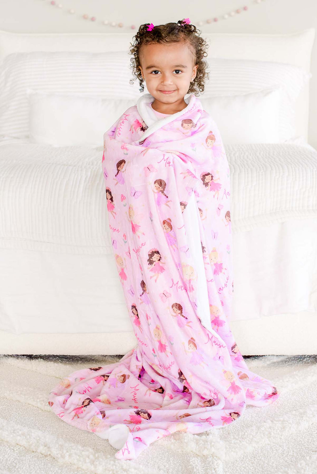 Toddler girl standing wrapped in the pink Fairy Wishes personalized kids' blanket with fairies and custom name print