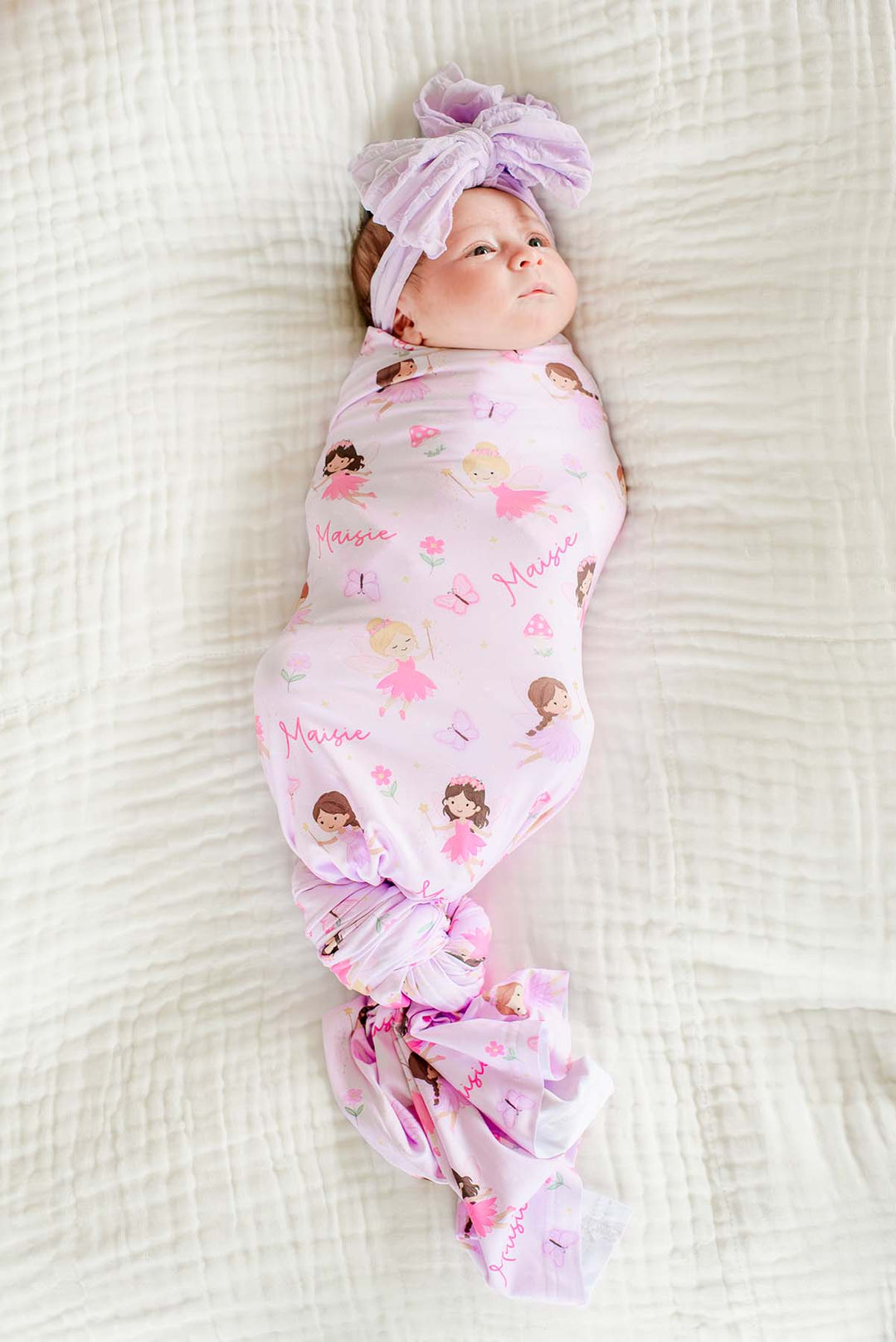 Baby wrapped in the pink Fairy Wishes personalized swaddle with fairies and custom name print, wearing a lavender bow headwrap