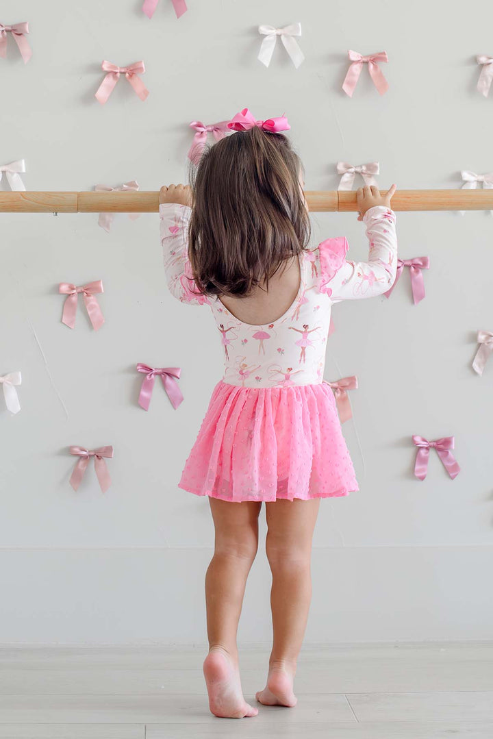 Back view of a girl on tiptoe at a ballet barre, wearing a Caden Lane scoop-back leotard in a ballerina print with a pink swiss dot mesh skirt, paired with a pink hair bow.