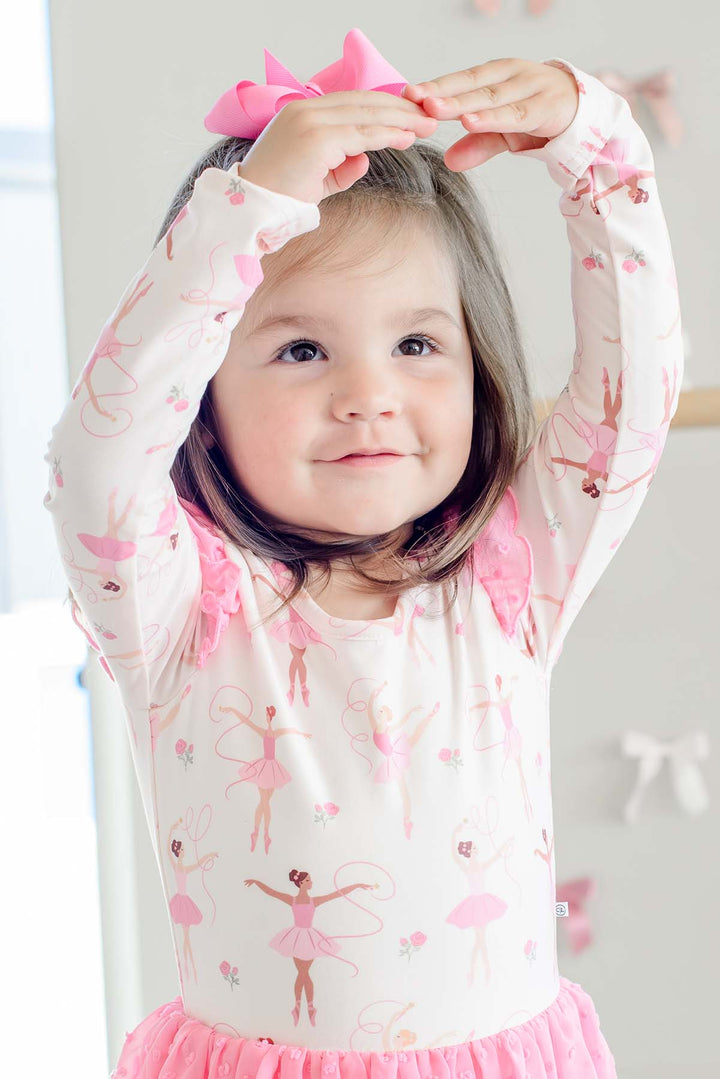 Close-up of a smiling girl with arms overhead in ballet pose, wearing a Caden Lane long-sleeve leotard in a ballerina print with pink flutter sleeves, paired with a pink hair bow.