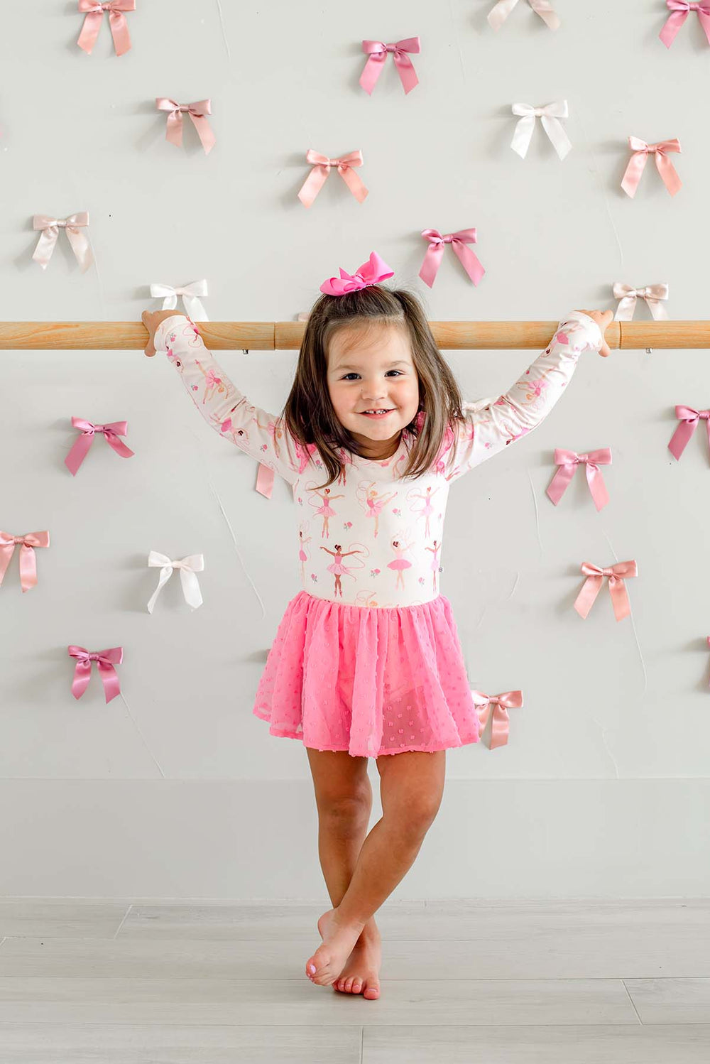 Smiling girl holding a ballet barre with crossed ankles, wearing a Caden Lane long-sleeve leotard in a ballerina print with a pink swiss dot mesh skirt.