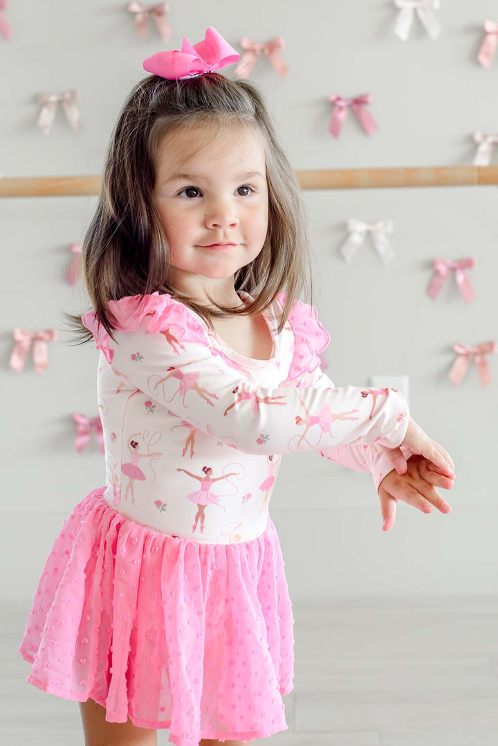 Girl dancing with arms extended in front of a ballet barre, wearing a Caden Lane ballerina print leotard with pink swiss dot flutter sleeves and skirt, paired with a pink hair bow.