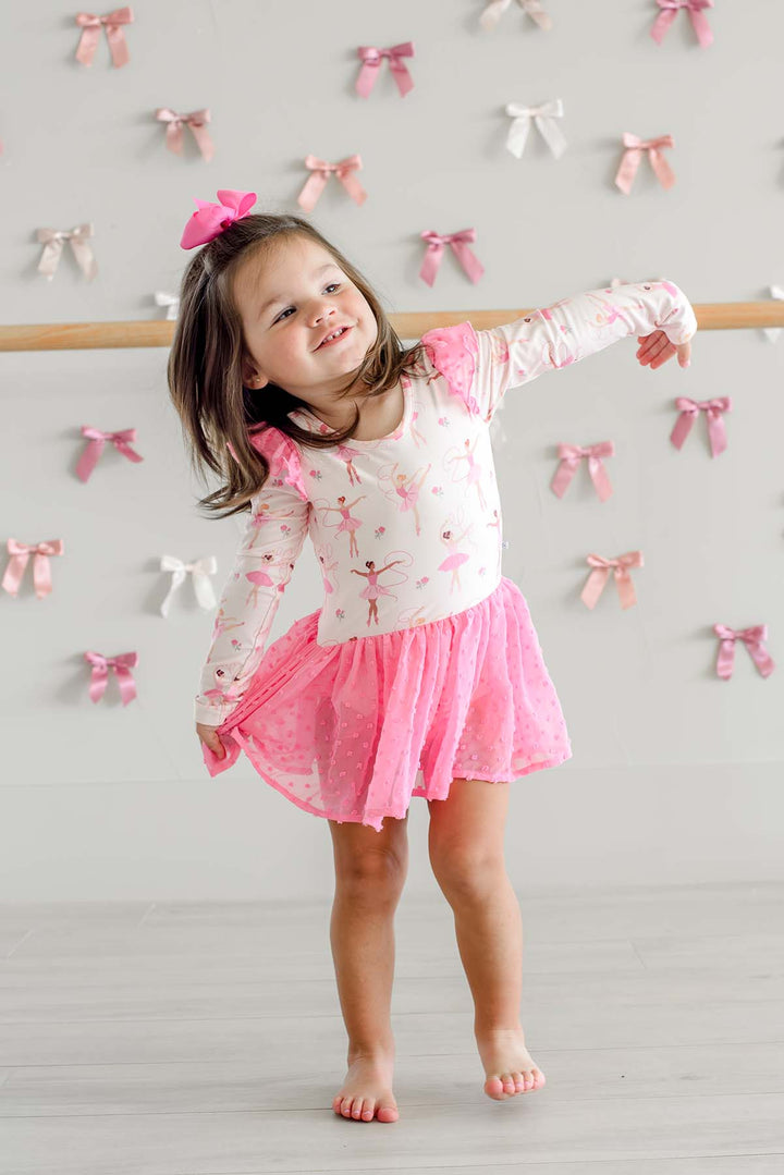 Smiling girl dancing with one arm out and holding her skirt, wearing a Caden Lane long-sleeve leotard in a ballerina print with a pink swiss dot mesh skirt.