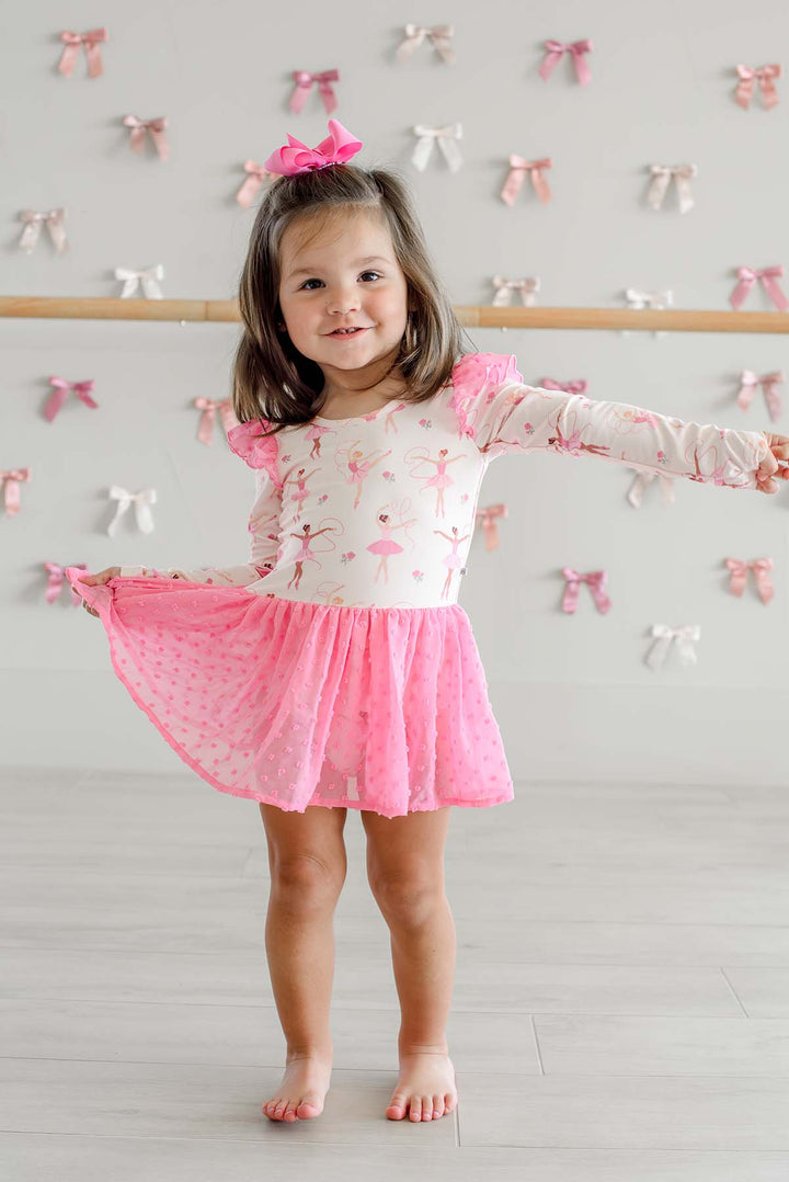 Girl holding out the pink swiss dot skirt of a Caden Lane long-sleeve ballerina print leotard while standing in front of a ballet barre and satin bow wall.