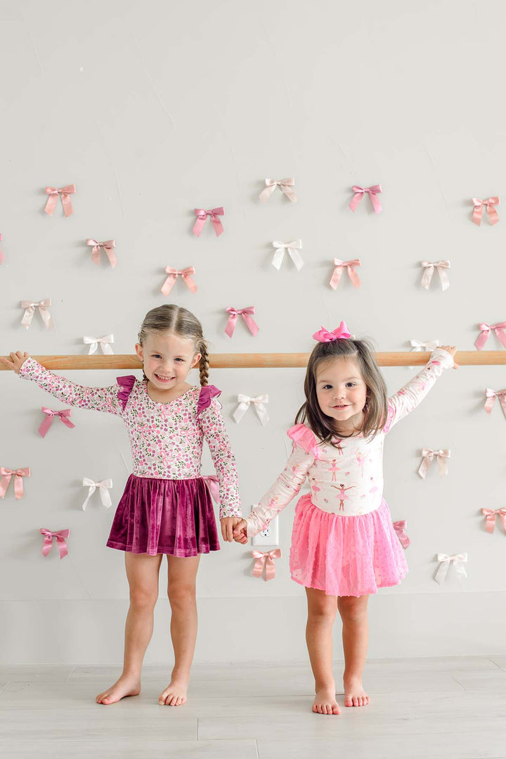 Two smiling girls holding hands at a ballet barre, one wearing a Caden Lane ballerina print leotard with a pink swiss dot skirt, beside a girl in a floral and bow print leotard with a berry velvet skirt.