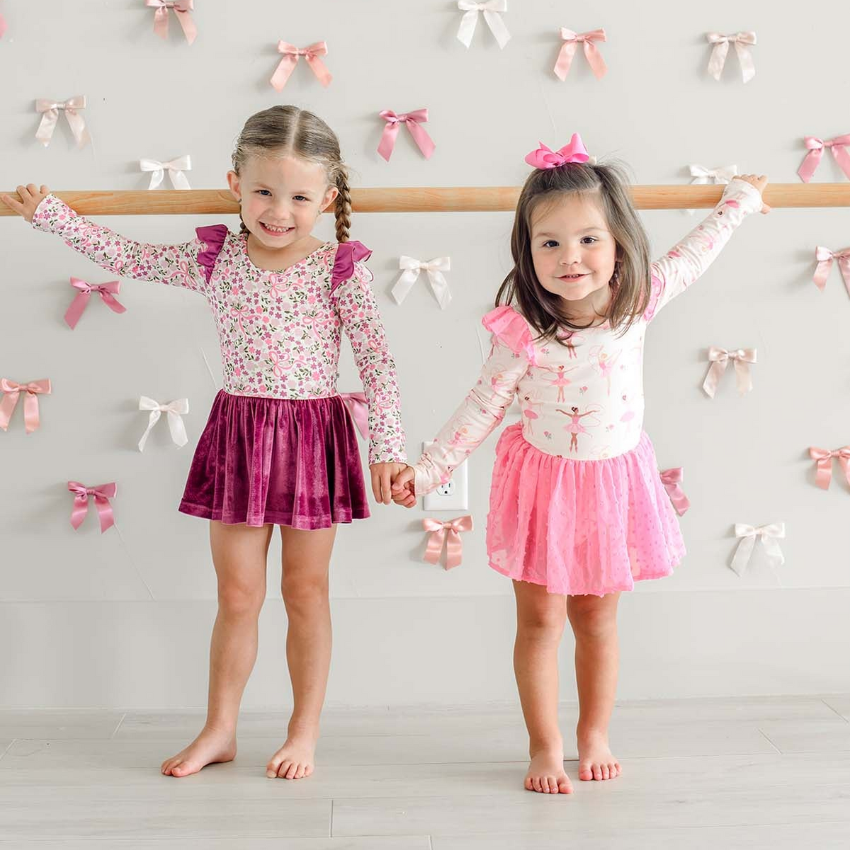 Two smiling girls holding a ballet barre, wearing Caden Lane long sleeve leotards in Woodland Garden and Pirouette Posies prints featuring florals, paired with velvet and tulle skirts.