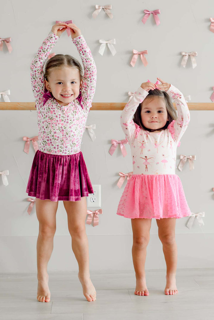 Two girls posing with arms overhead in front of a ballet barre and satin bow wall, one wearing a Caden Lane long-sleeve leotard in a pink floral and bow print with a berry velvet skirt, beside a girl in a ballerina print leotard with a pink tulle skirt.