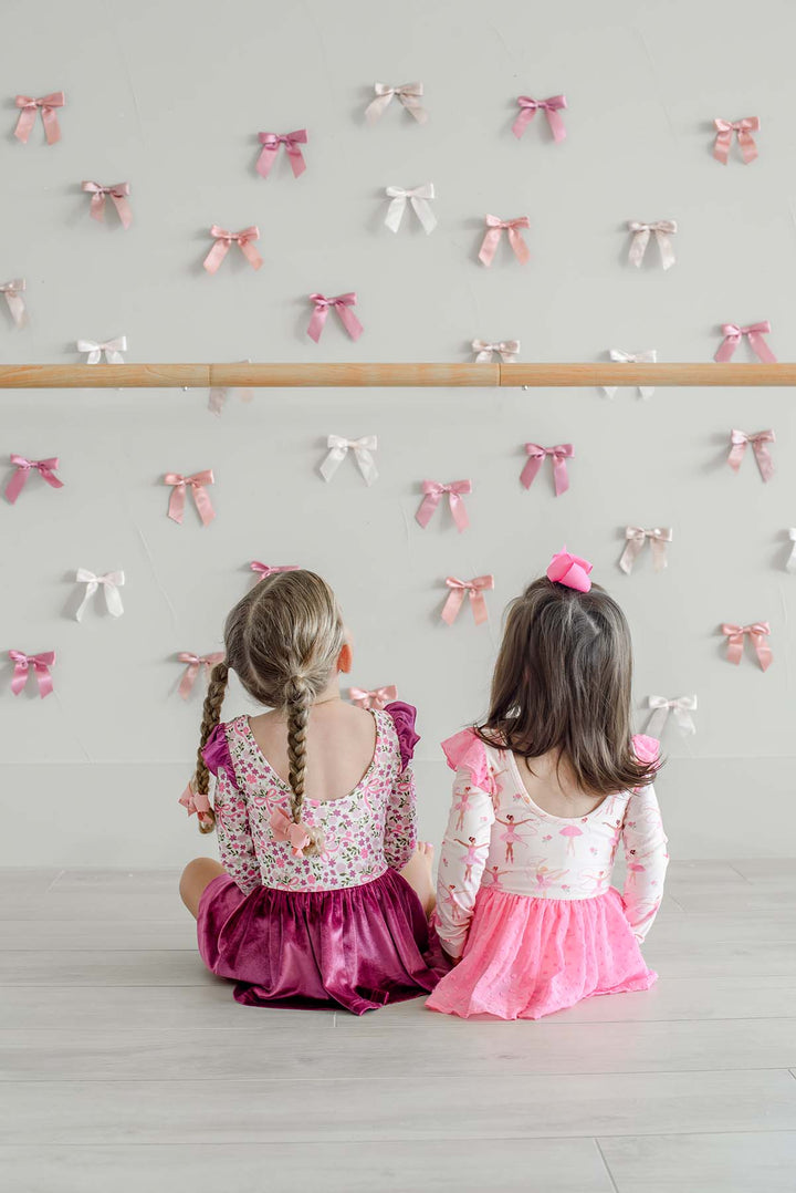 Back view of two girls sitting in front of a ballet barre and satin bow wall, one in a Caden Lane scoop-back ballerina print leotard with a pink swiss dot skirt, beside a girl in a floral and bow print leotard.