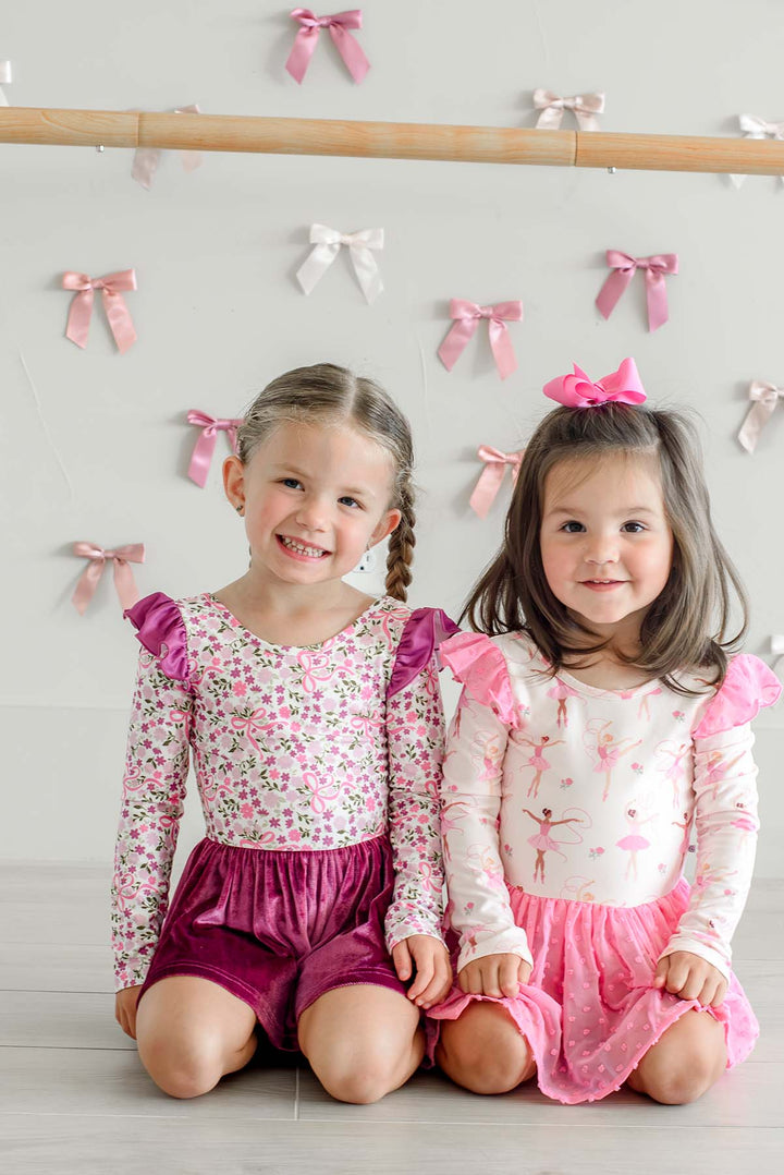 Two smiling girls kneeling in front of a ballet barre, one in a Caden Lane floral and bow print leotard with berry flutter sleeves and a velvet skirt, the other in a ballerina print leotard with a pink tulle skirt.