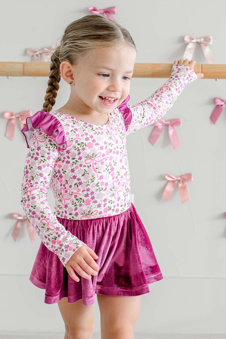 Smiling girl with a braid holding a ballet barre, wearing a Caden Lane long-sleeve leotard in a pink floral and bow print with berry flutter sleeves and a berry velvet skirt.