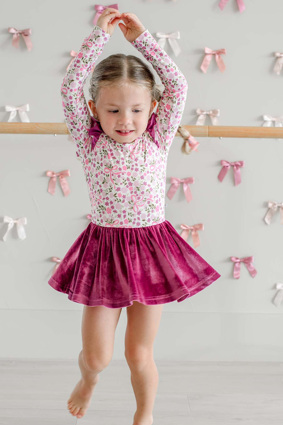 Girl hopping with arms overhead in front of a ballet barre, wearing a Caden Lane long-sleeve leotard in a pink floral and bow print with a twirling berry velvet skirt.