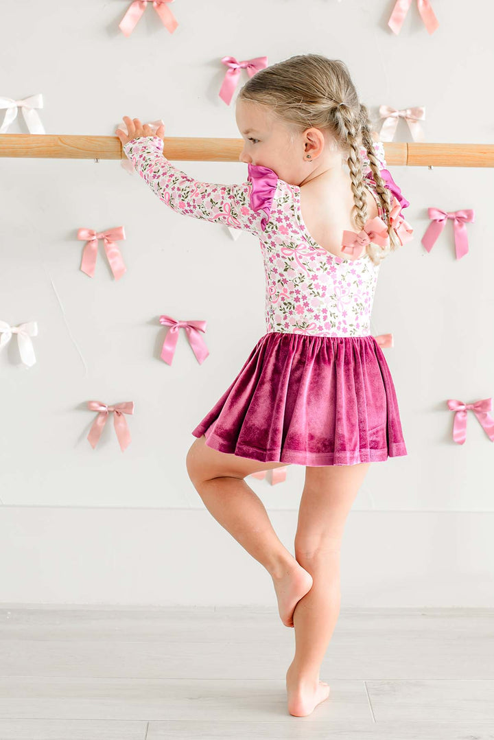 Back view of a girl in passé pose at a ballet barre, wearing a Caden Lane scoop-back leotard in a pink floral and bow print with a berry velvet skirt.