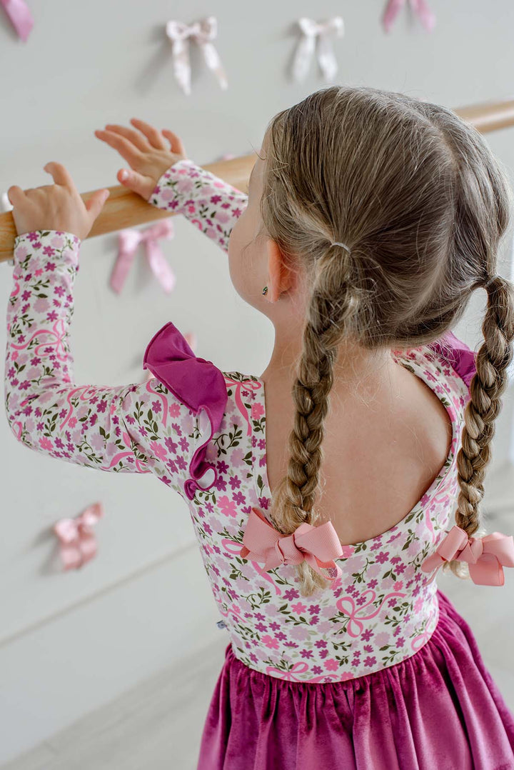 Close-up back view of a girl with braided pigtails holding a ballet barre, showing the scoop back and berry flutter sleeves of a Caden Lane floral and bow print leotard.