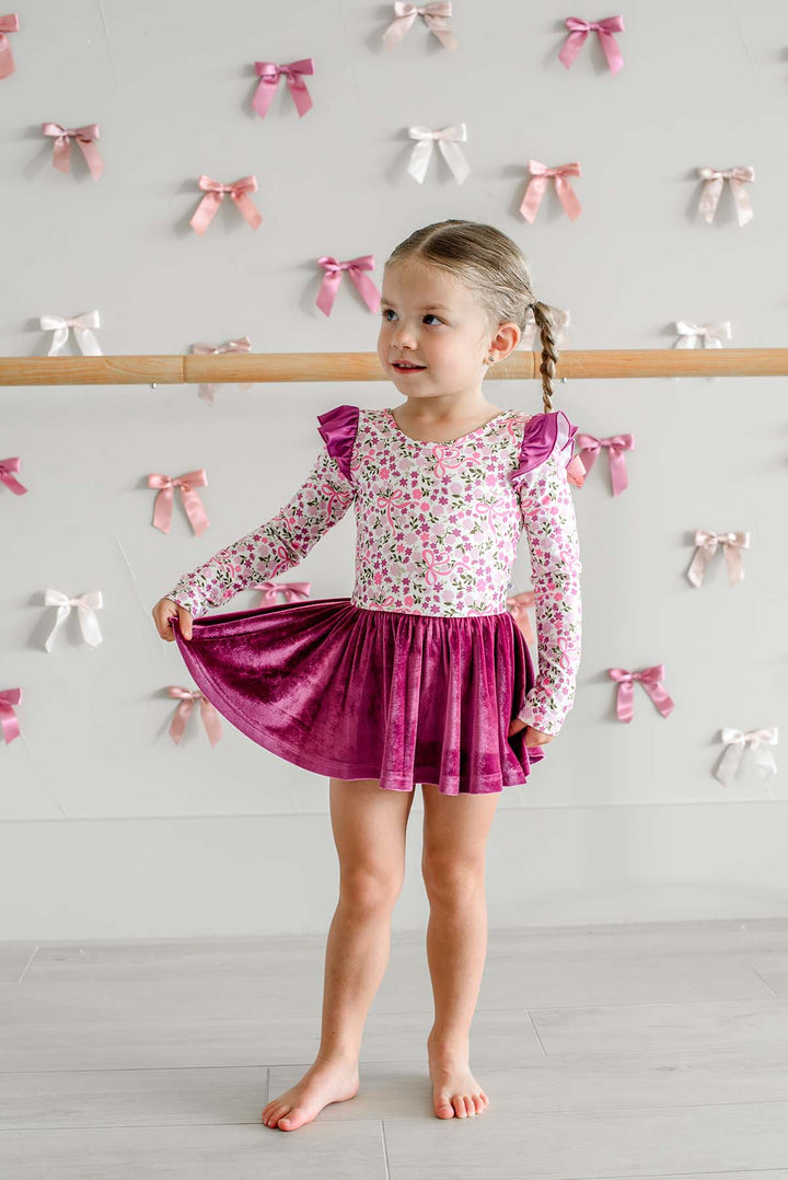 Girl standing in front of a satin bow wall holding out the berry velvet skirt of a Caden Lane long-sleeve leotard in a pink floral and bow print with flutter sleeves.