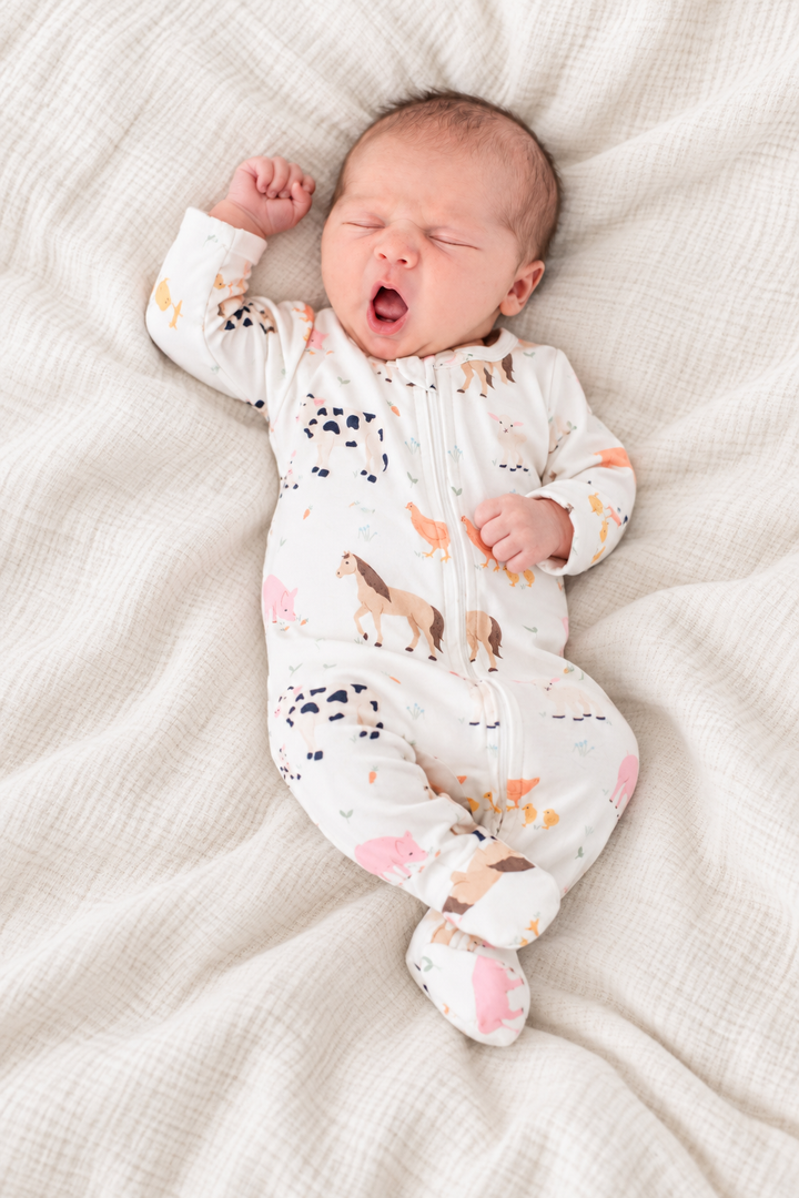 Yawning newborn in Caden Lane farm animal print zip-up footie pajamas stretching on a blanket