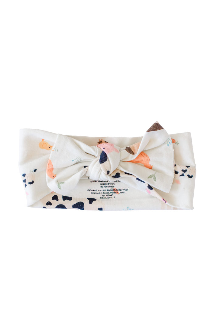 A Caden Lane fabric headband with a large bow, featuring a white base with a colorful animal print and small black spots, shown on a plain white background.