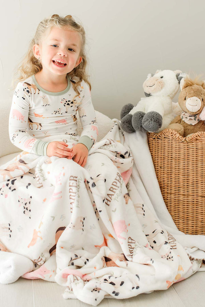 Toddler girl in Caden Lane farm animal pajamas sitting with a personalized 'Harper Peyton' minky blanket beside a basket of stuffed animals