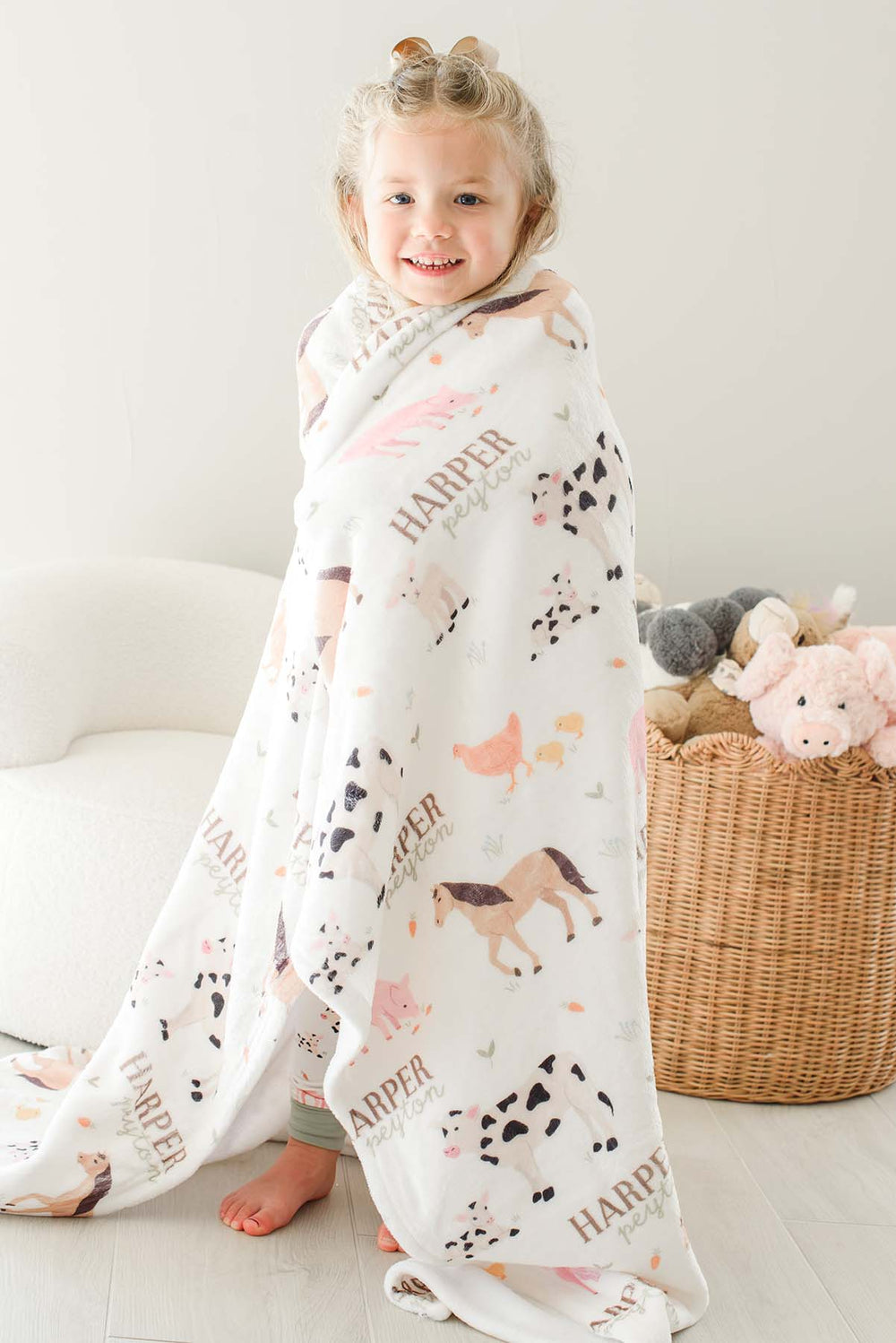 Smiling toddler girl wrapped in Caden Lane personalized 'Harper Peyton' farm animal minky blanket standing near a basket of stuffed animals