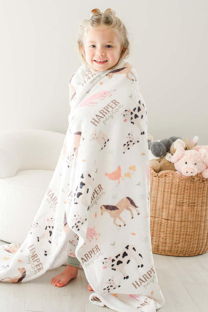 Smiling toddler girl wrapped in Caden Lane personalized 'Harper Peyton' farm animal minky blanket standing near a basket of stuffed animals