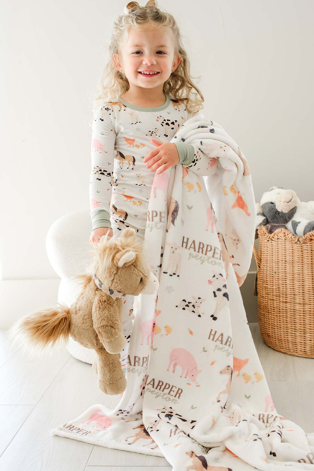 Smiling toddler girl in Caden Lane farm animal pajamas holding a personalized 'Harper Peyton' minky blanket and stuffed horse toy